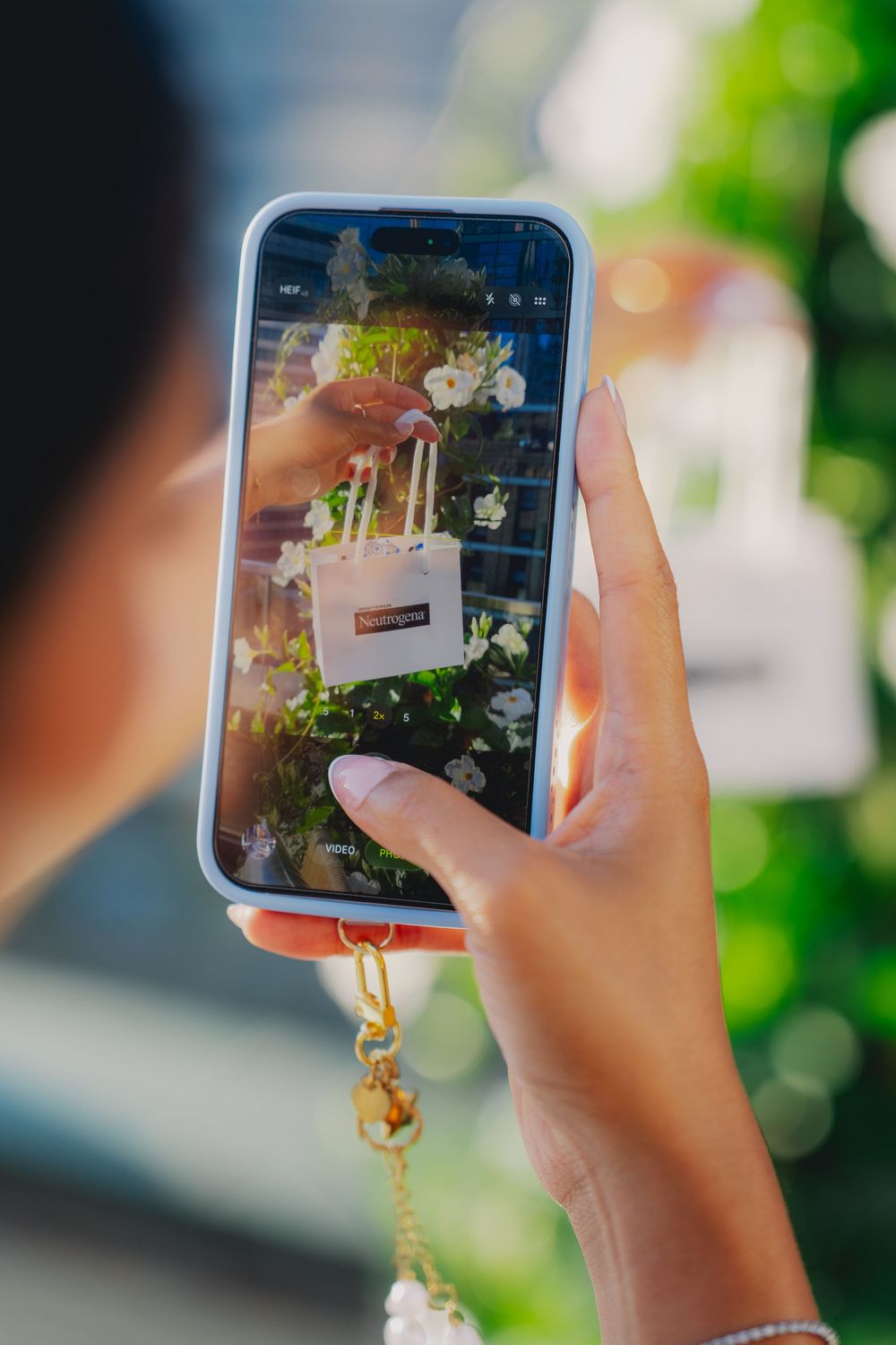 Woman photographing a Neutrogena shopping bag with white flowers on her smartphone outdoors.