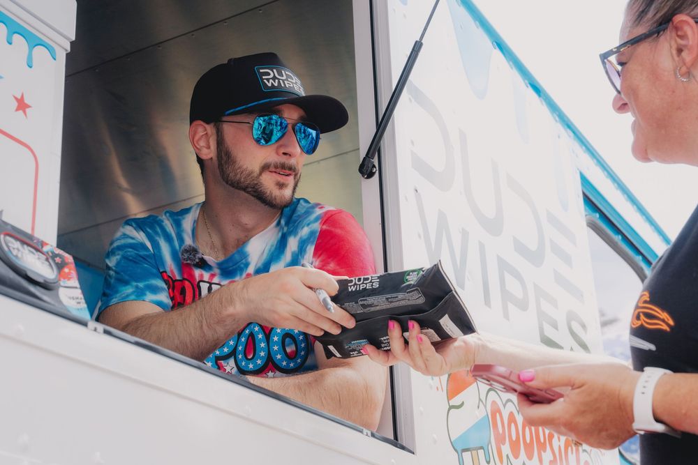 Man in sunglasses and tie-dye shirt handing items from a food truck window at an outdoor event.