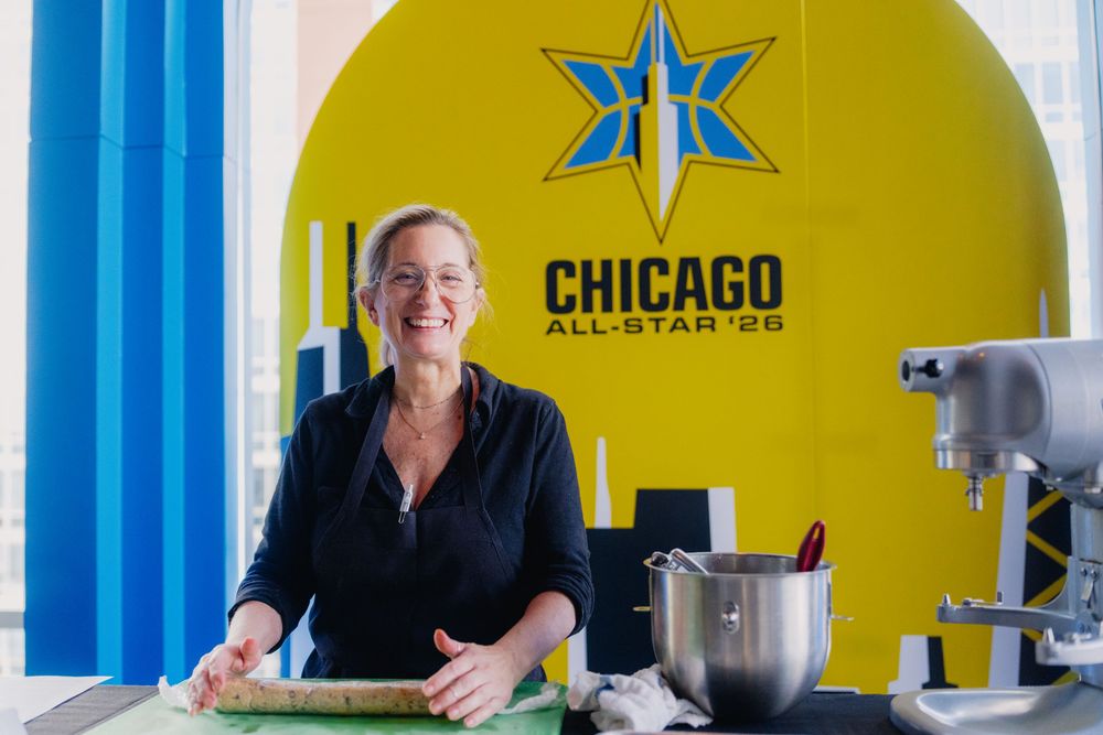 Woman smiling at a baking station with a Chicago All-Star 26 logo backdrop in a bright, modern kitchen.