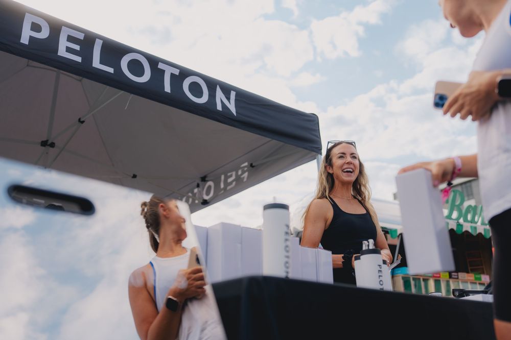 Two women smiling at a Peloton outdoor event booth on a sunny day.