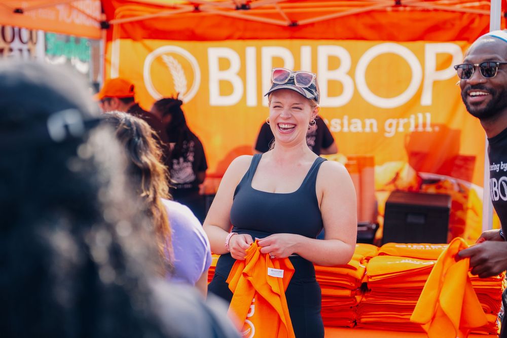 Woman smiling at BIBIBOP Asian Grill event, handing out orange shirts at an outdoor festival booth.