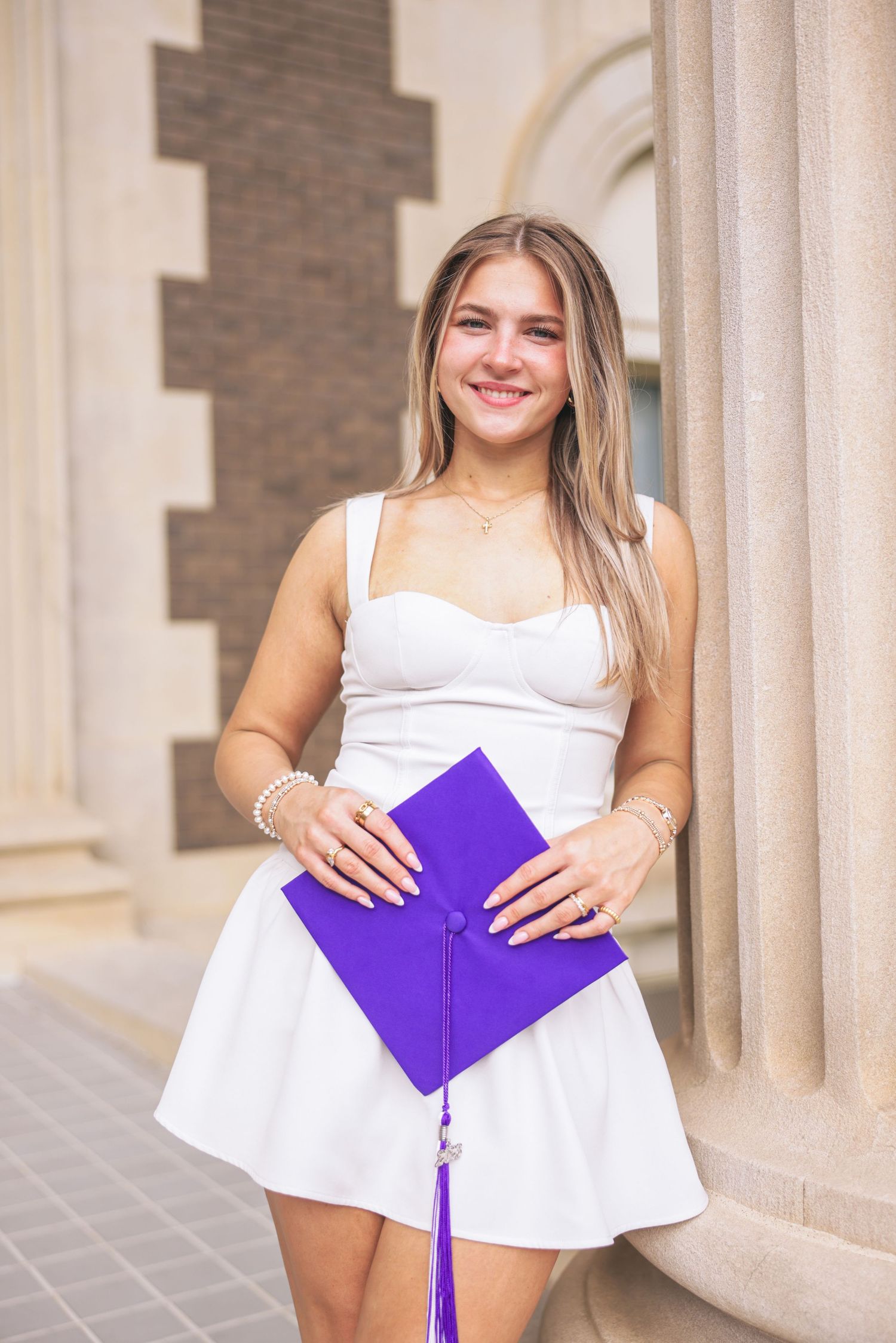 Young woman with curly hair smiling in white dress and black graduation gown drape outside a building.