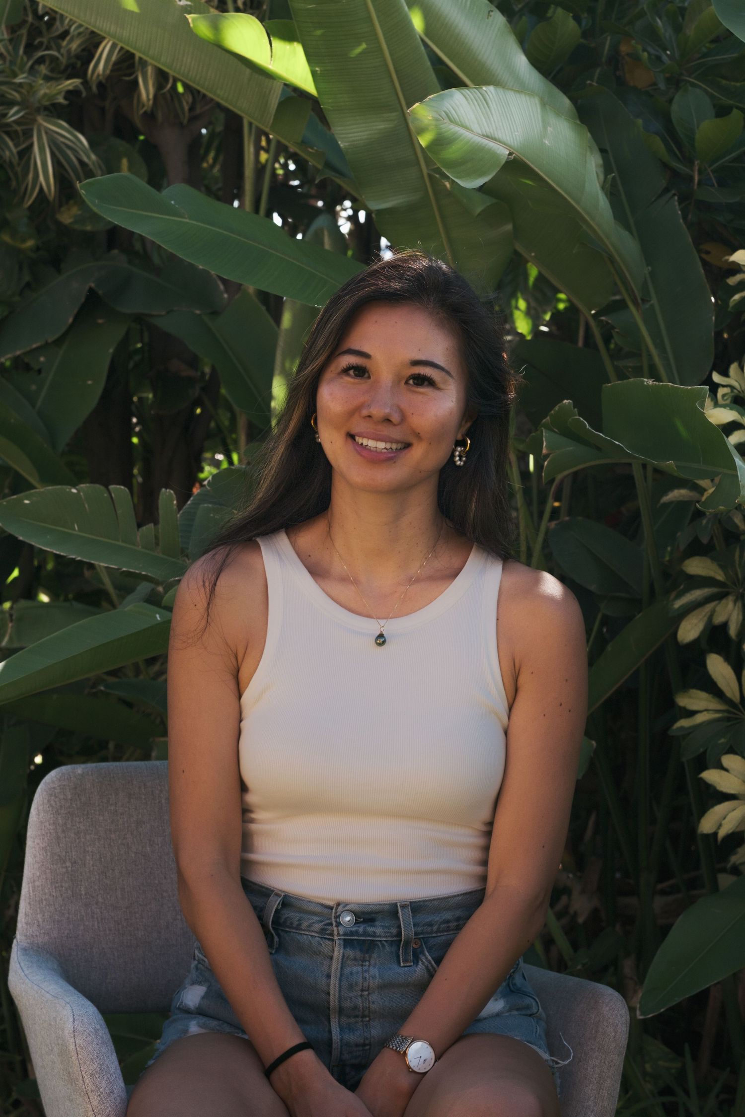 Young woman smiling in a white tank top and denim shorts, seated outdoors with lush tropical green leaves behind her.