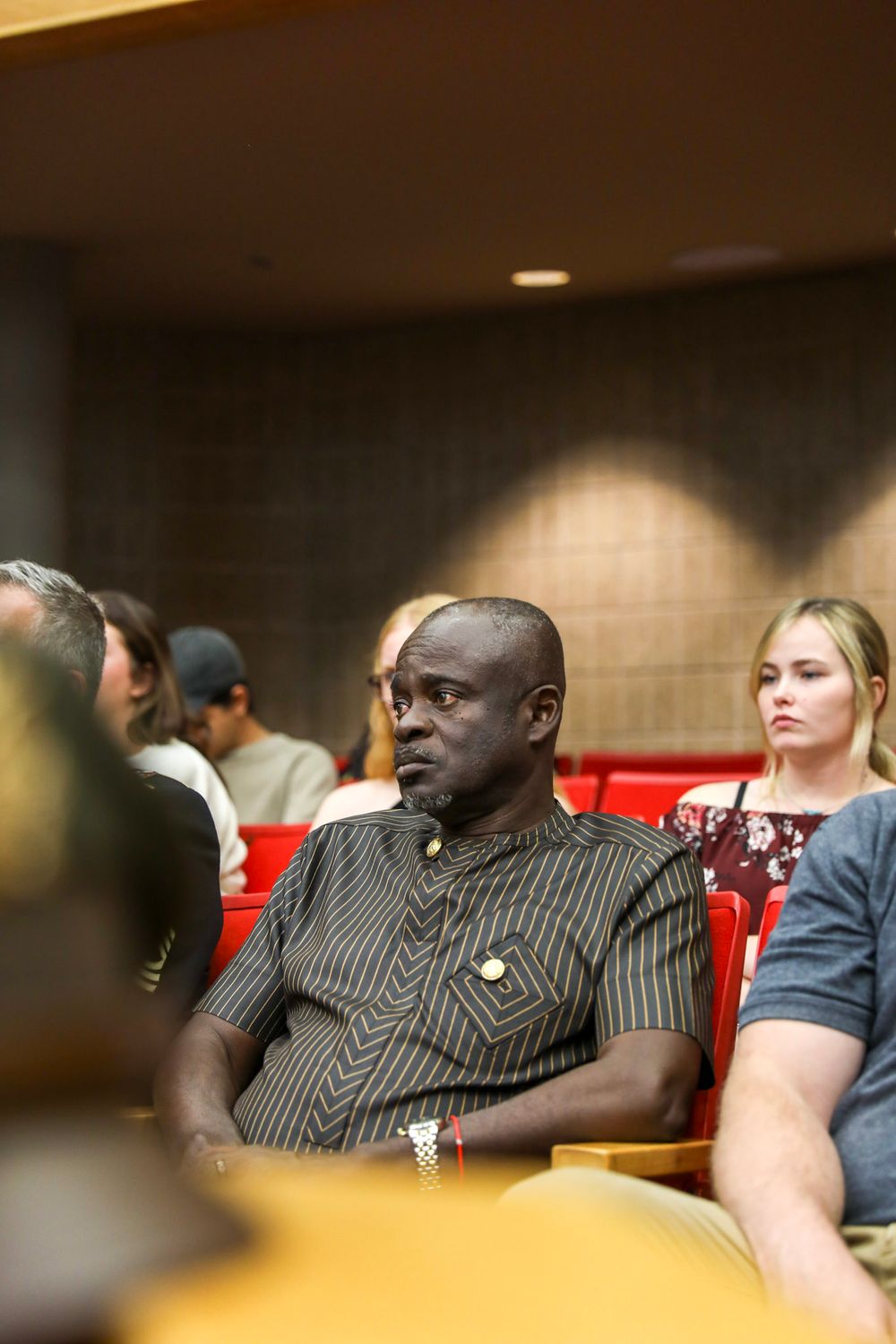 Man in striped shirt sitting attentively in red auditorium seats among a diverse audience.