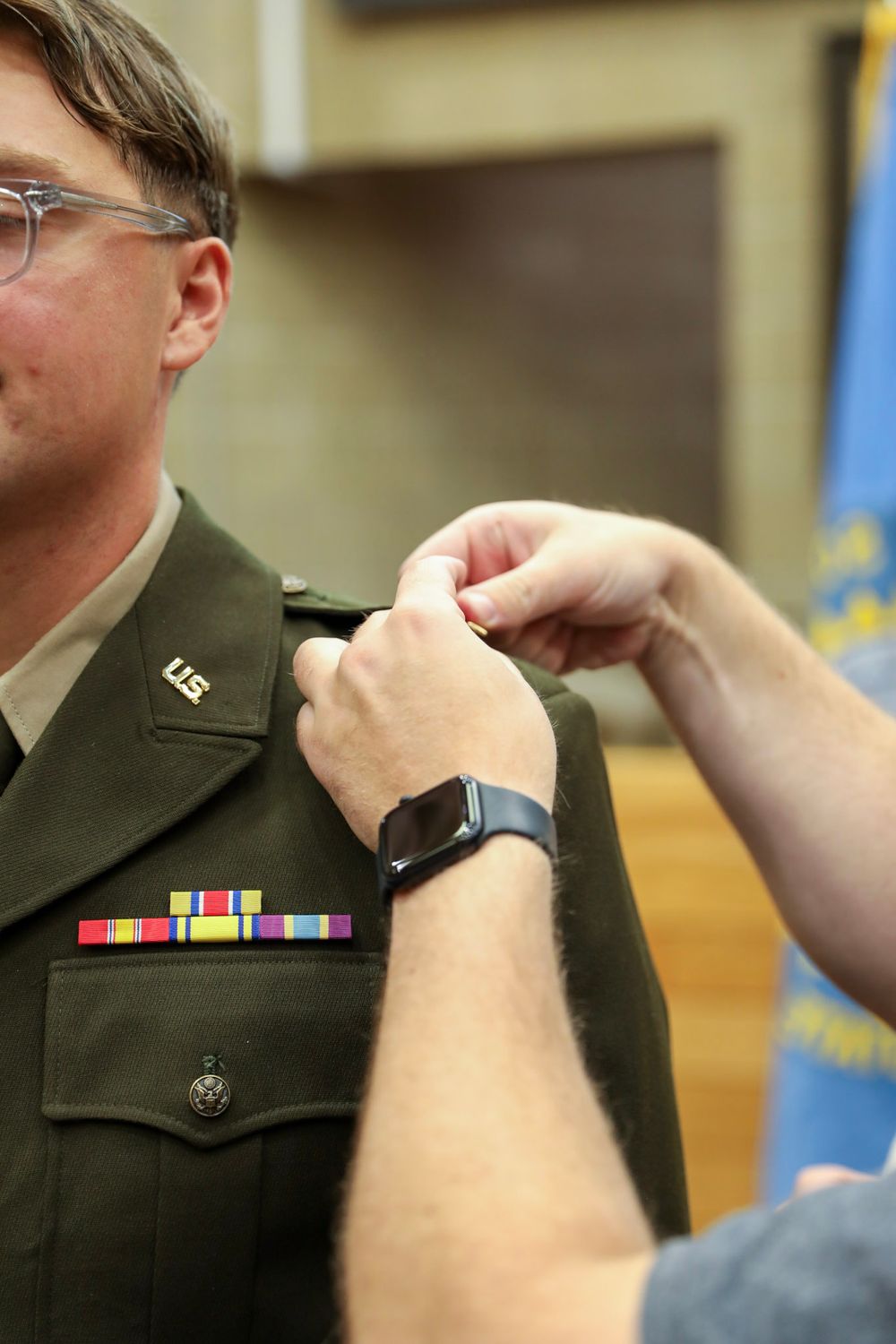 Military officer in dress uniform receiving a medal pinning ceremony, wearing ribbons and U.S. insignia on olive green jacket.