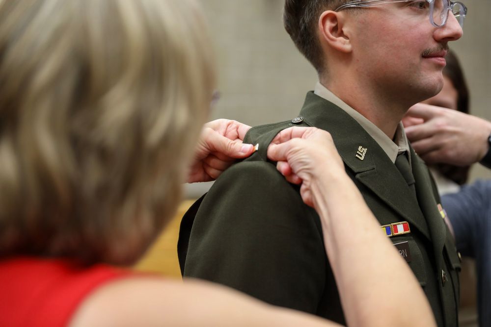Woman pinning rank insignia on military officer's olive green dress uniform during a formal promotion ceremony.