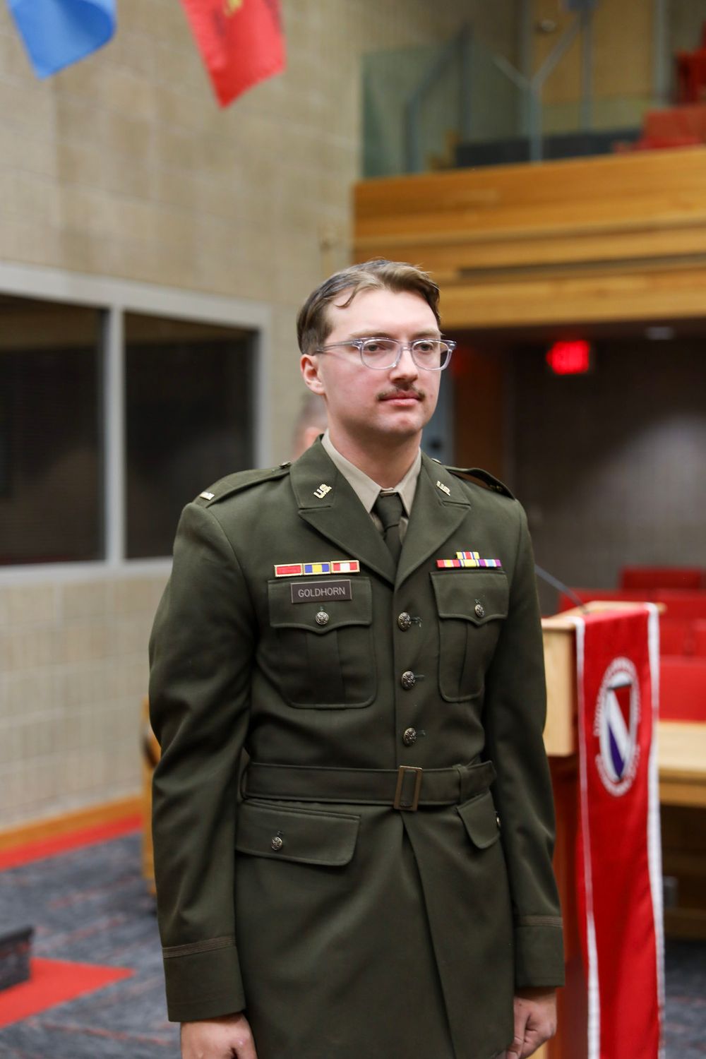 Young man in olive green military dress uniform with ribbons and medals stands at attention indoors near flags.