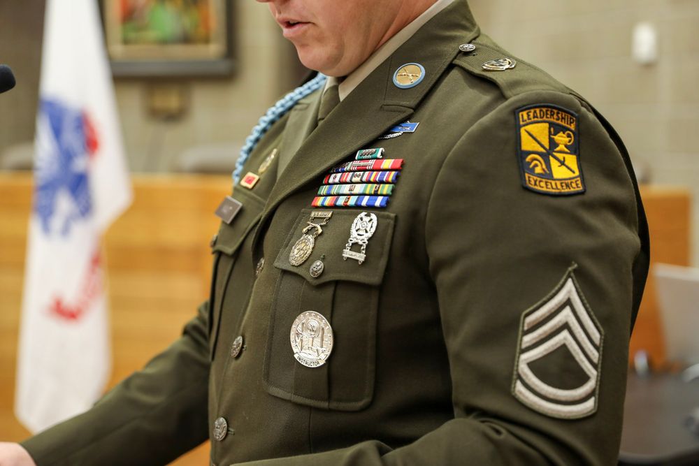 U.S. Army Staff Sergeant in dress green uniform displaying medals, ribbons, and unit patches at a formal ceremony.