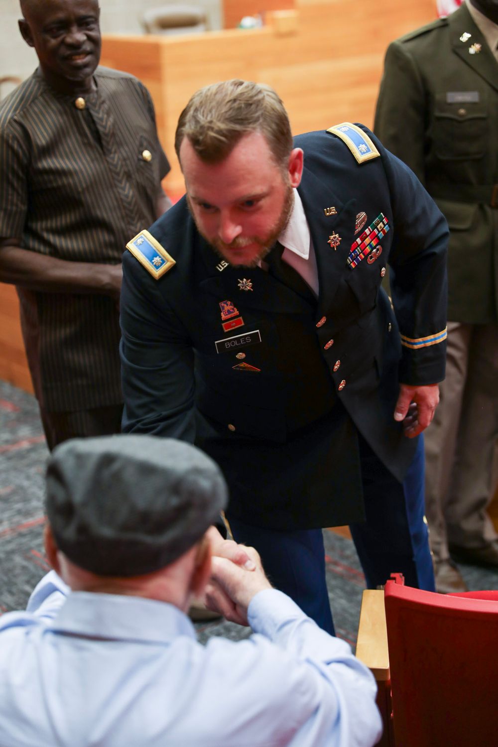 Military officer in dress uniform shaking hands with a seated person at a formal ceremony.