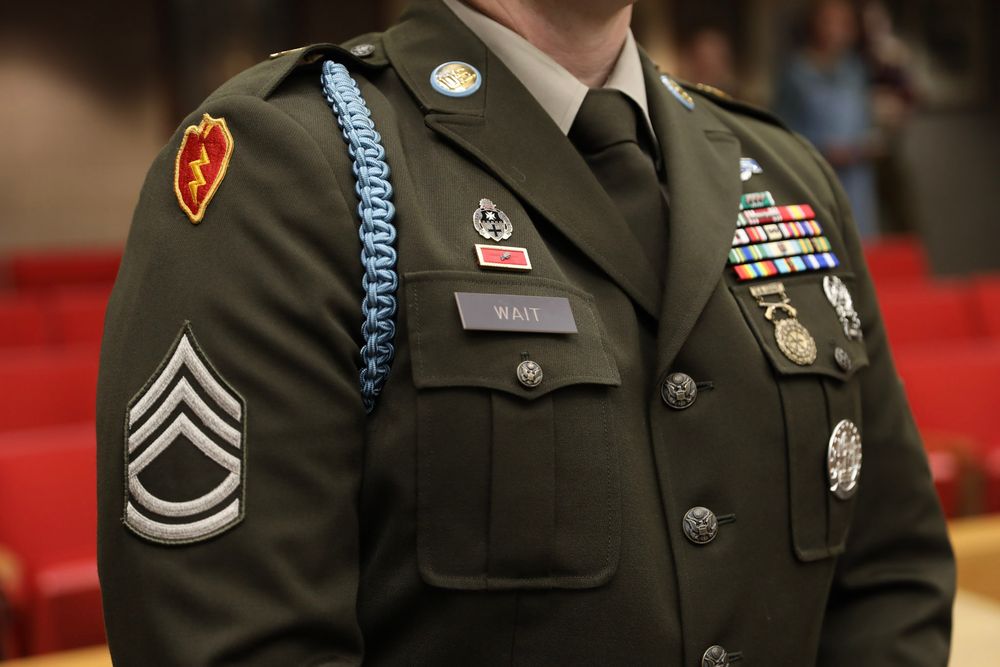 US Army Staff Sergeant in dress green uniform with medals, ribbons, and blue cord on display.