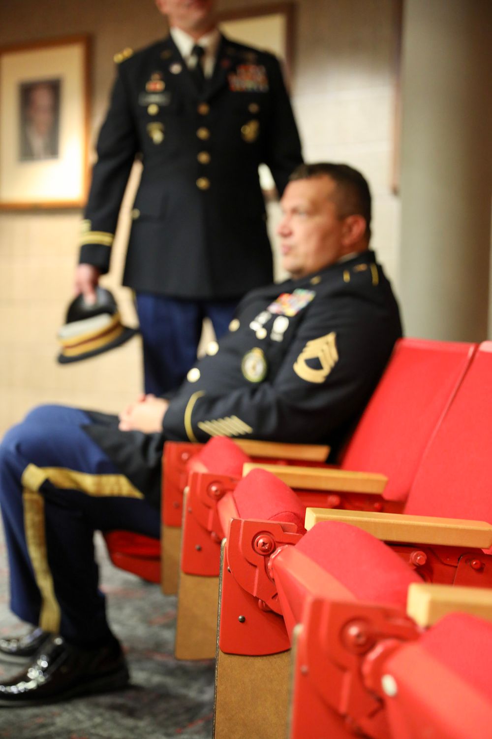 Two U.S. Army soldiers in dress uniforms sit and stand in red auditorium chairs during a military ceremony.