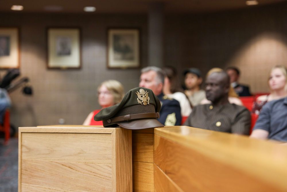 Military dress cap resting on wooden podium in formal meeting room with uniformed personnel seated in background.