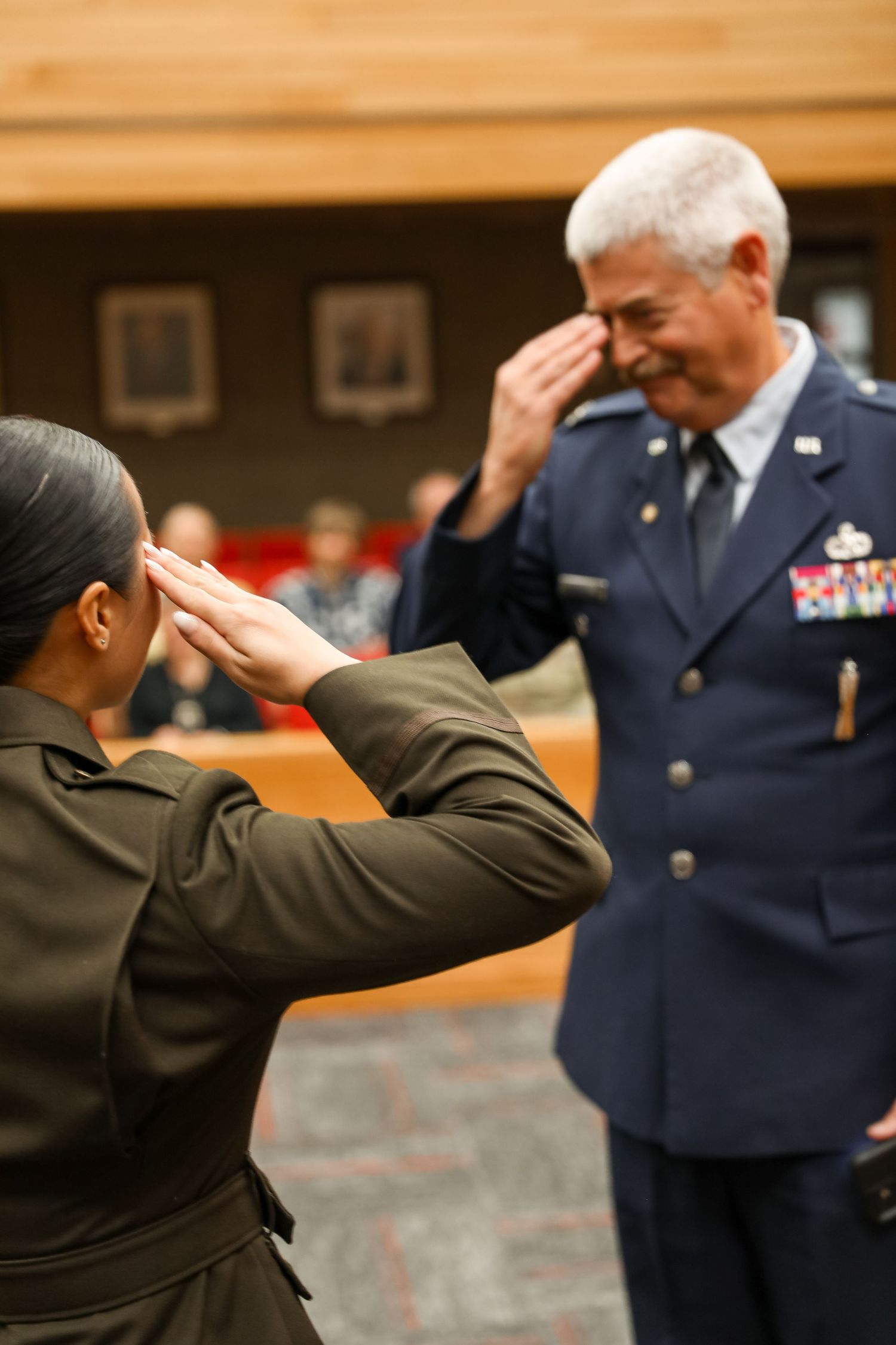 Two military officers in uniform exchanging salutes during a formal ceremony indoors.