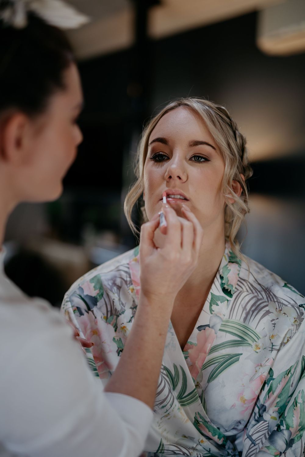 Professional makeup artist applying lipstick to someone wearing a tropical print robe during wedding preparations.