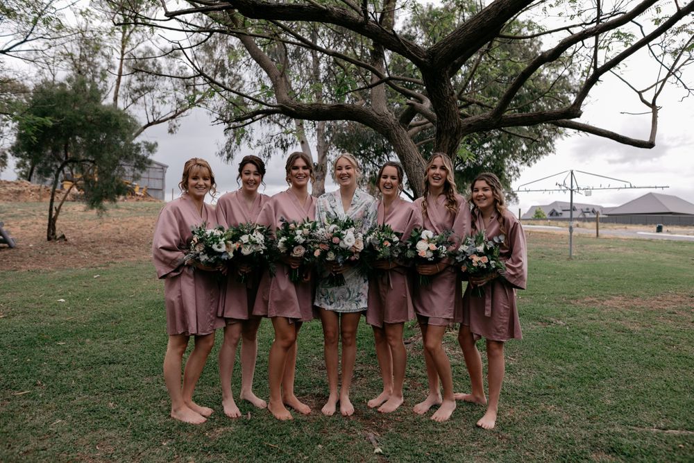 Bridal party in matching pink robes and bouquets poses under a large tree outdoors.