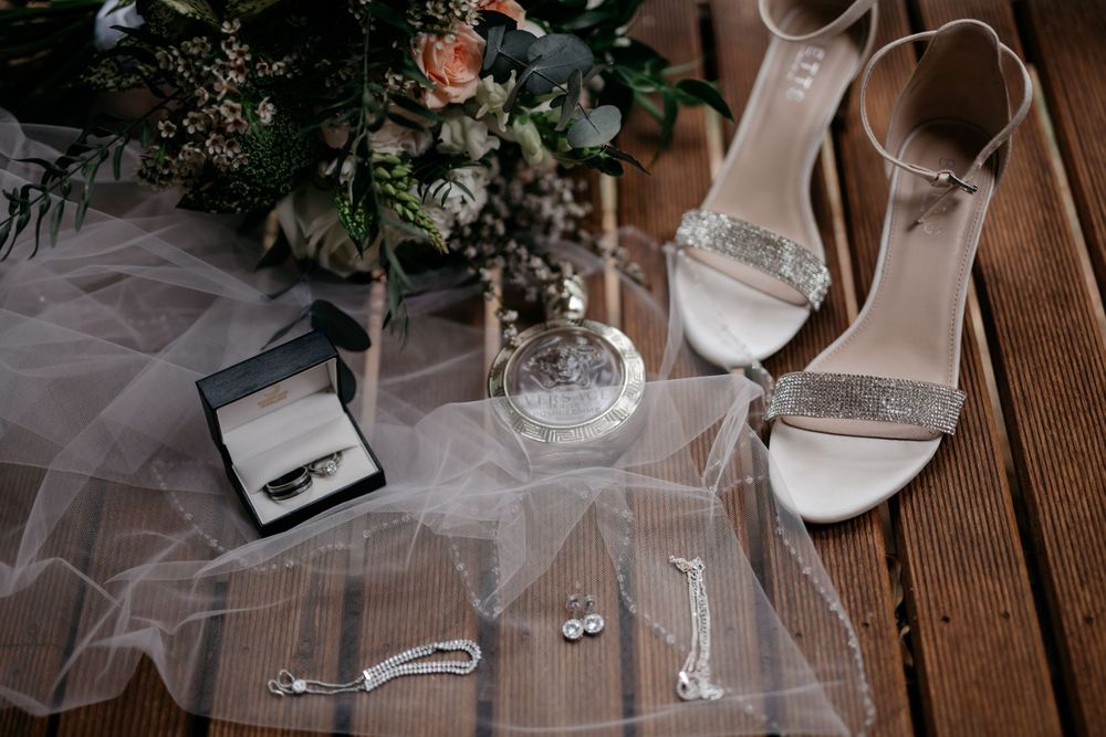 Wedding detail flatlay with white shoes, veil, jewelry and floral arrangements on wooden surface.