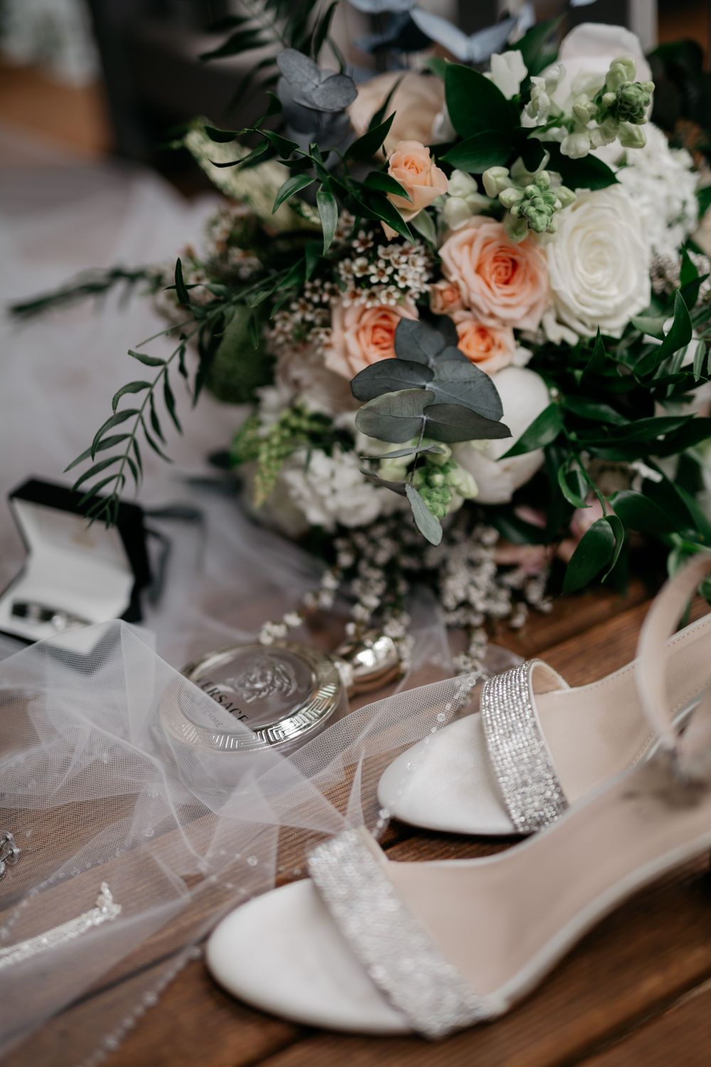 Wedding accessories including white heels, veil, and peachy floral bouquet arranged on wooden surface.