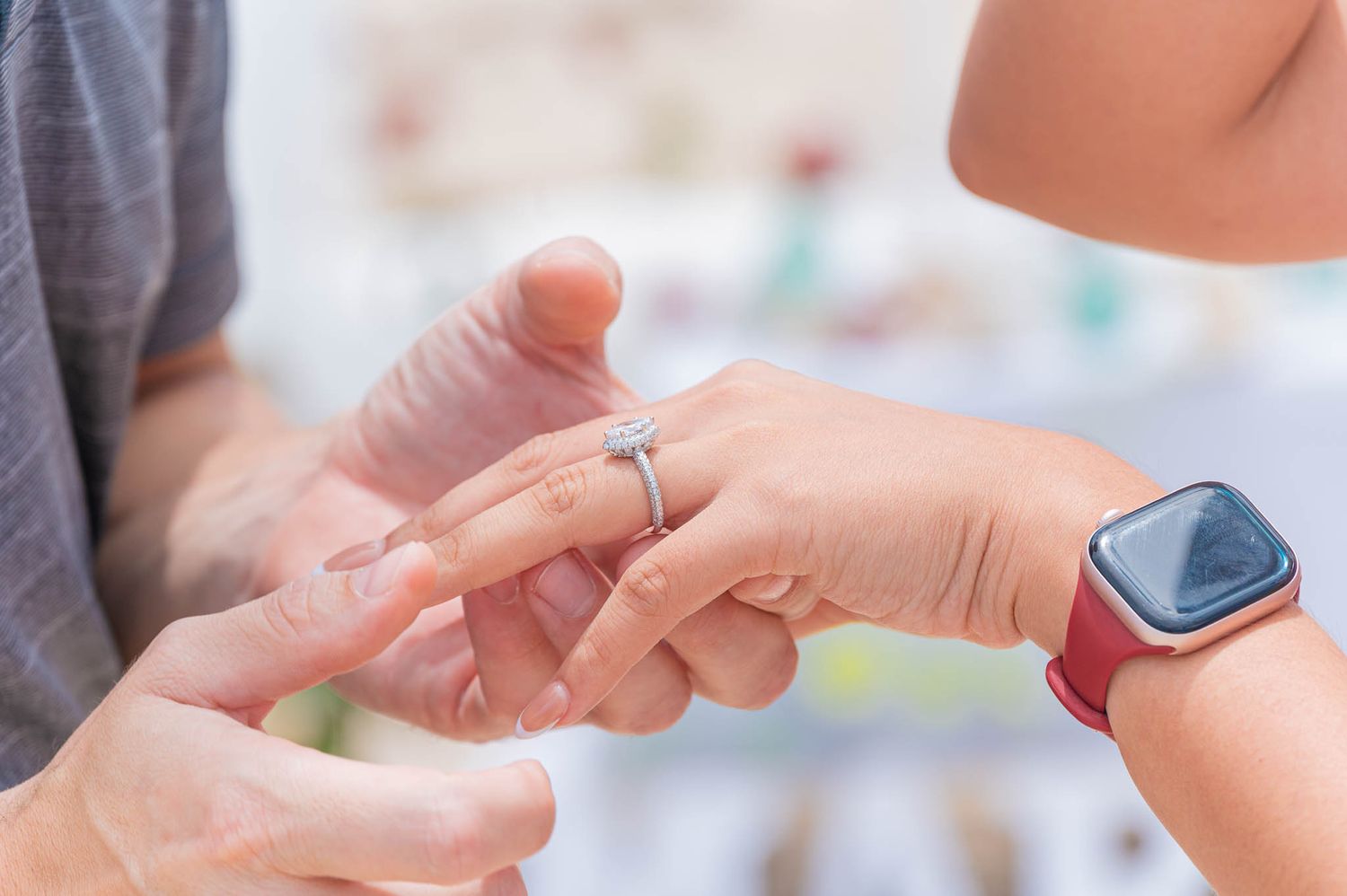 A person placing a diamond engagement ring on a partner's finger, with a red Apple Watch visible on the wrist.