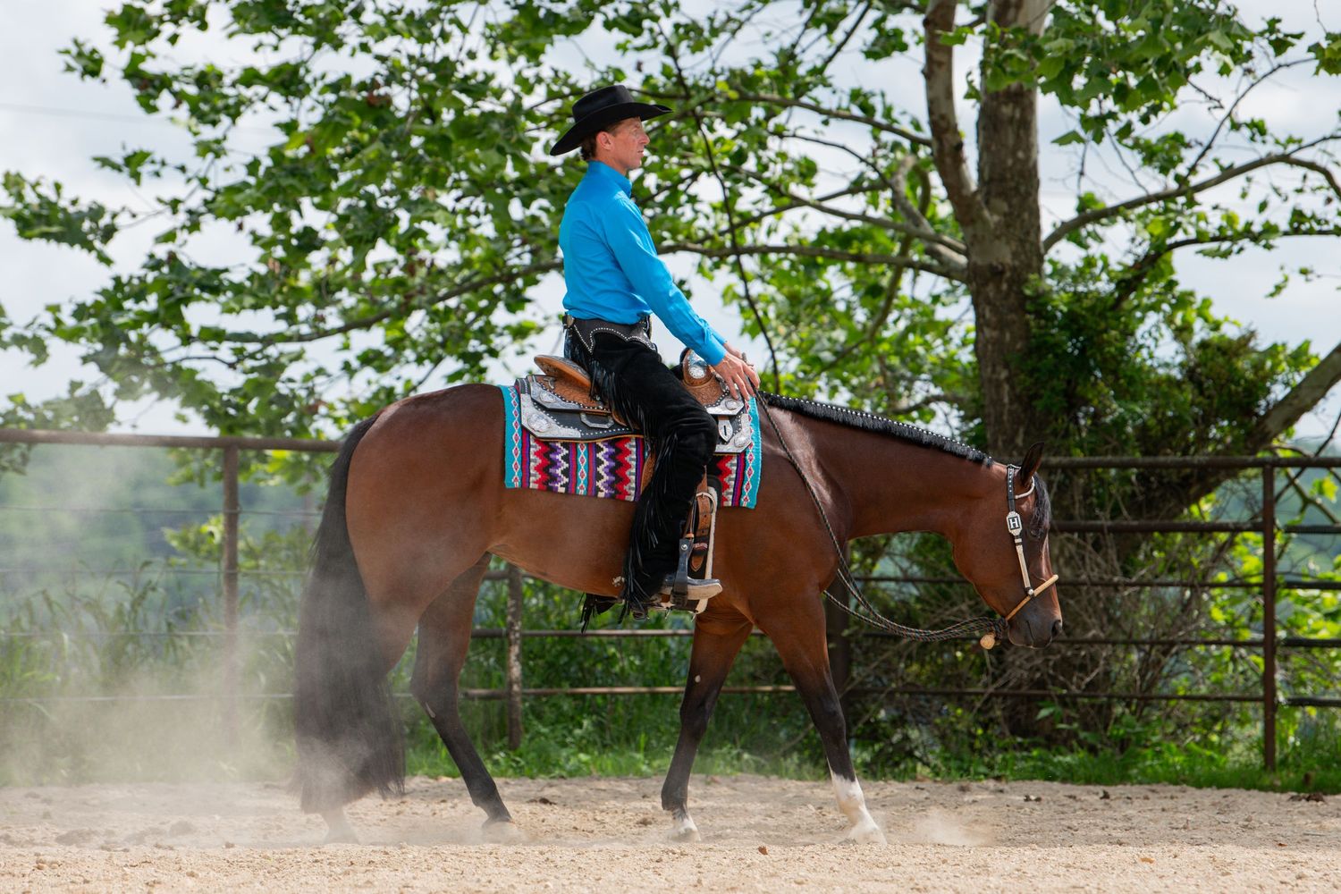 A rider in a blue shirt and black cowboy hat rides a bay horse in a sandy outdoor arena with green hills behind.