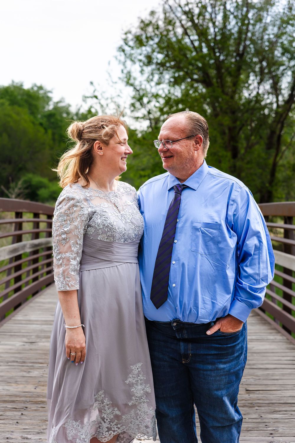 A smiling couple stands on a wooden bridge, woman in lace dress, man in blue shirt and tie, surrounded by green trees.