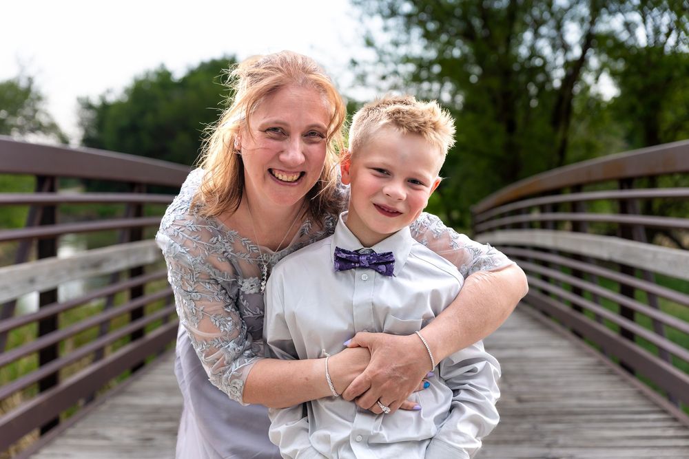 A smiling woman hugs a young boy in a bow tie on a wooden bridge outdoors.