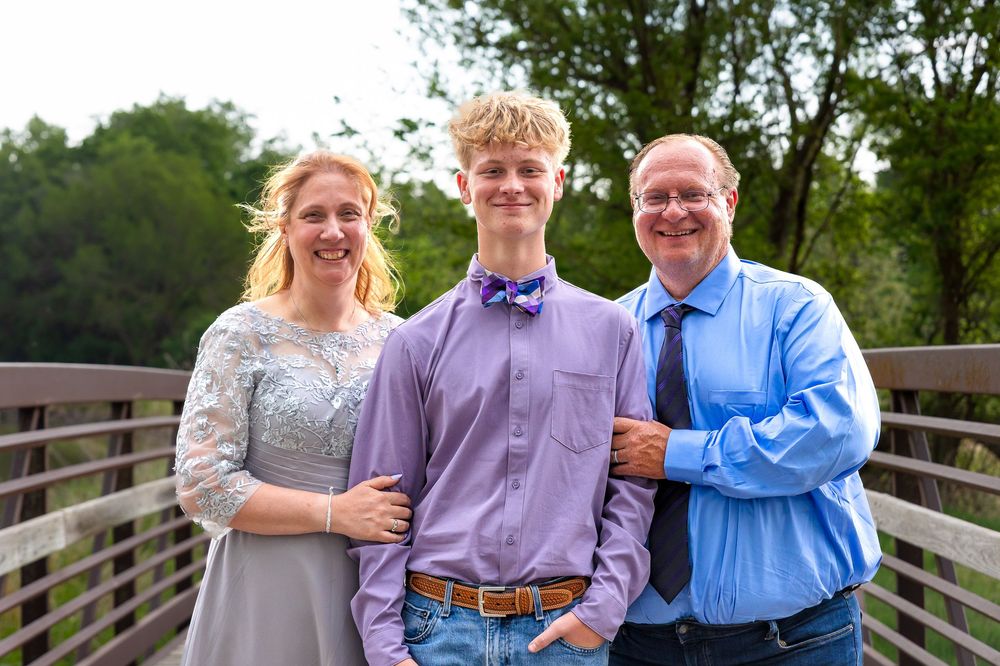 A family of three smiles together on a bridge, dressed in formal attire, surrounded by lush green trees.
