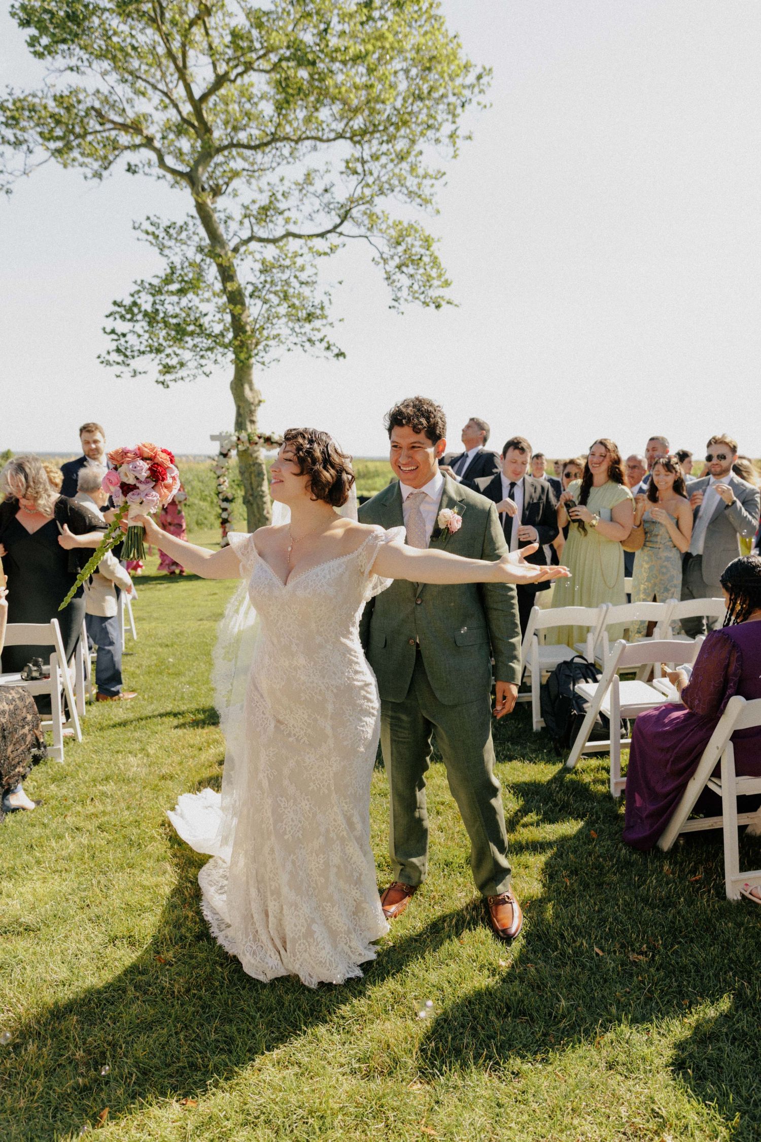 Joyful bride and groom walk back down the aisle after outdoor wedding ceremony on sunny green lawn.