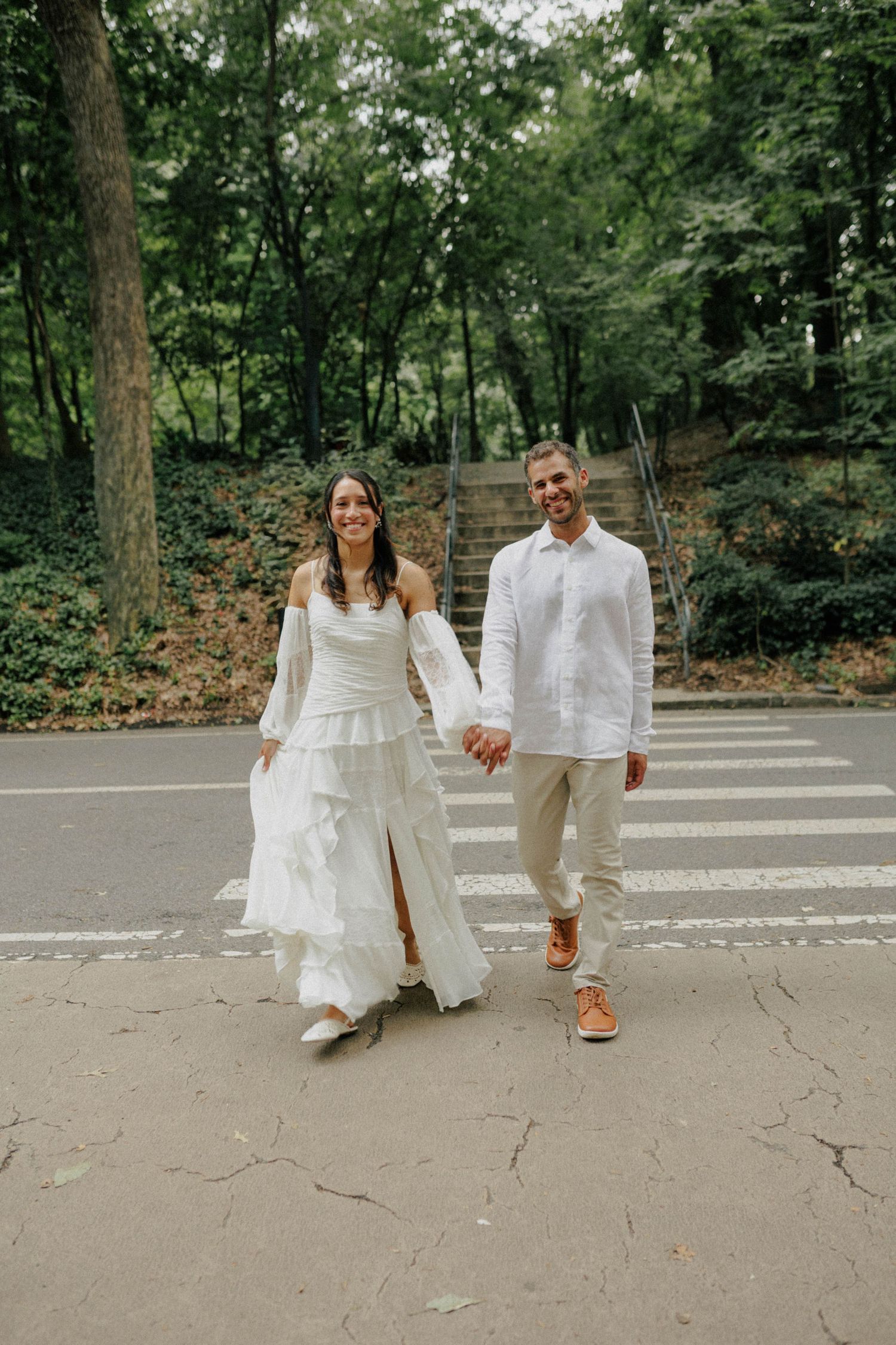 Newlywed couple holding hands, crossing a zebra crossing in a lush park, bride in white gown, groom in white shirt.