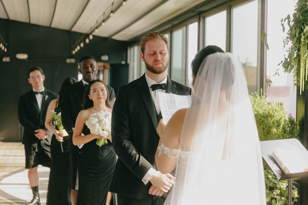 Groom in black tuxedo holding bride's hands during intimate indoor wedding ceremony.