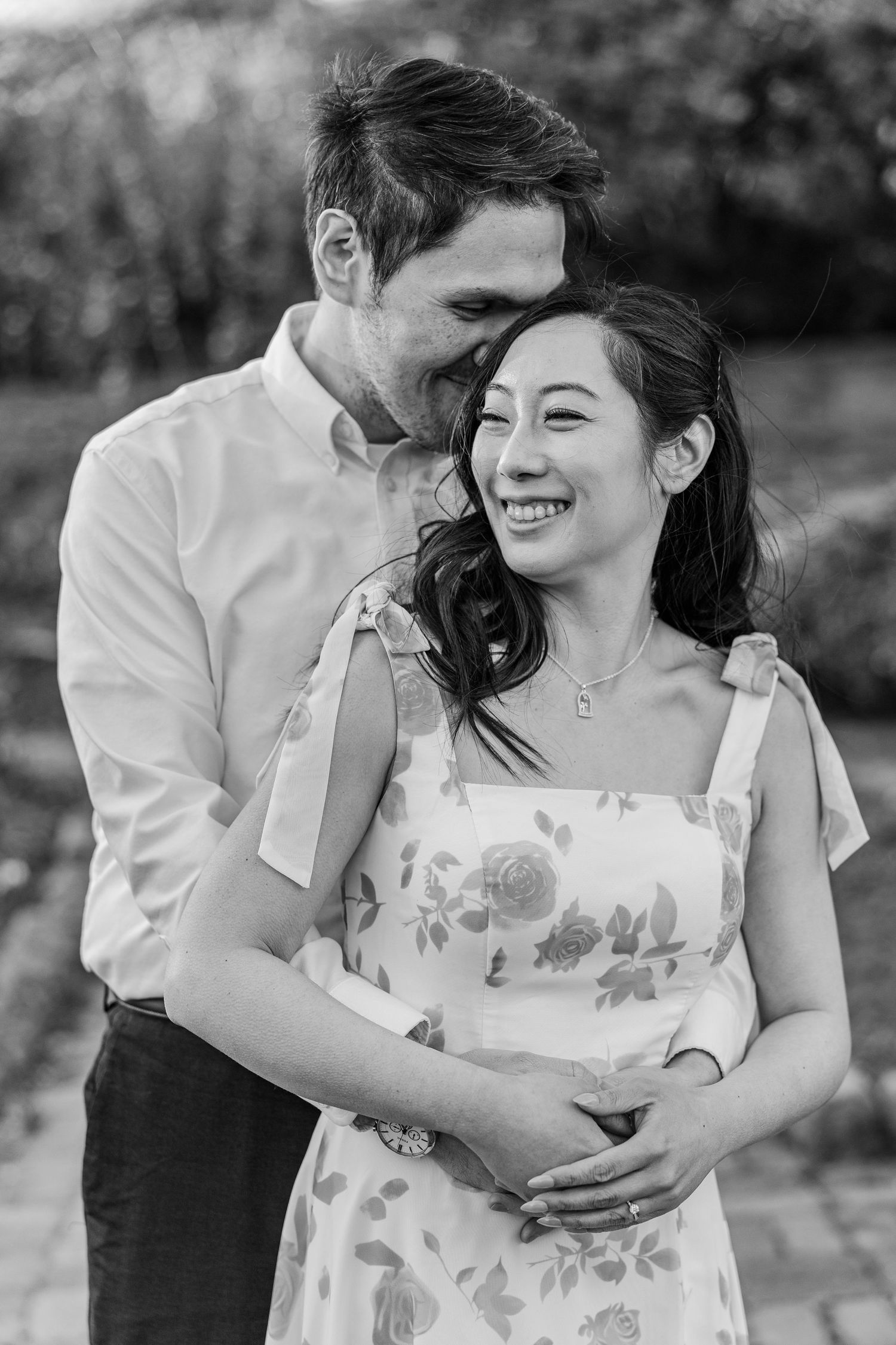 Black and white photo of a smiling couple embracing outdoors, woman in floral dress, man in light shirt.