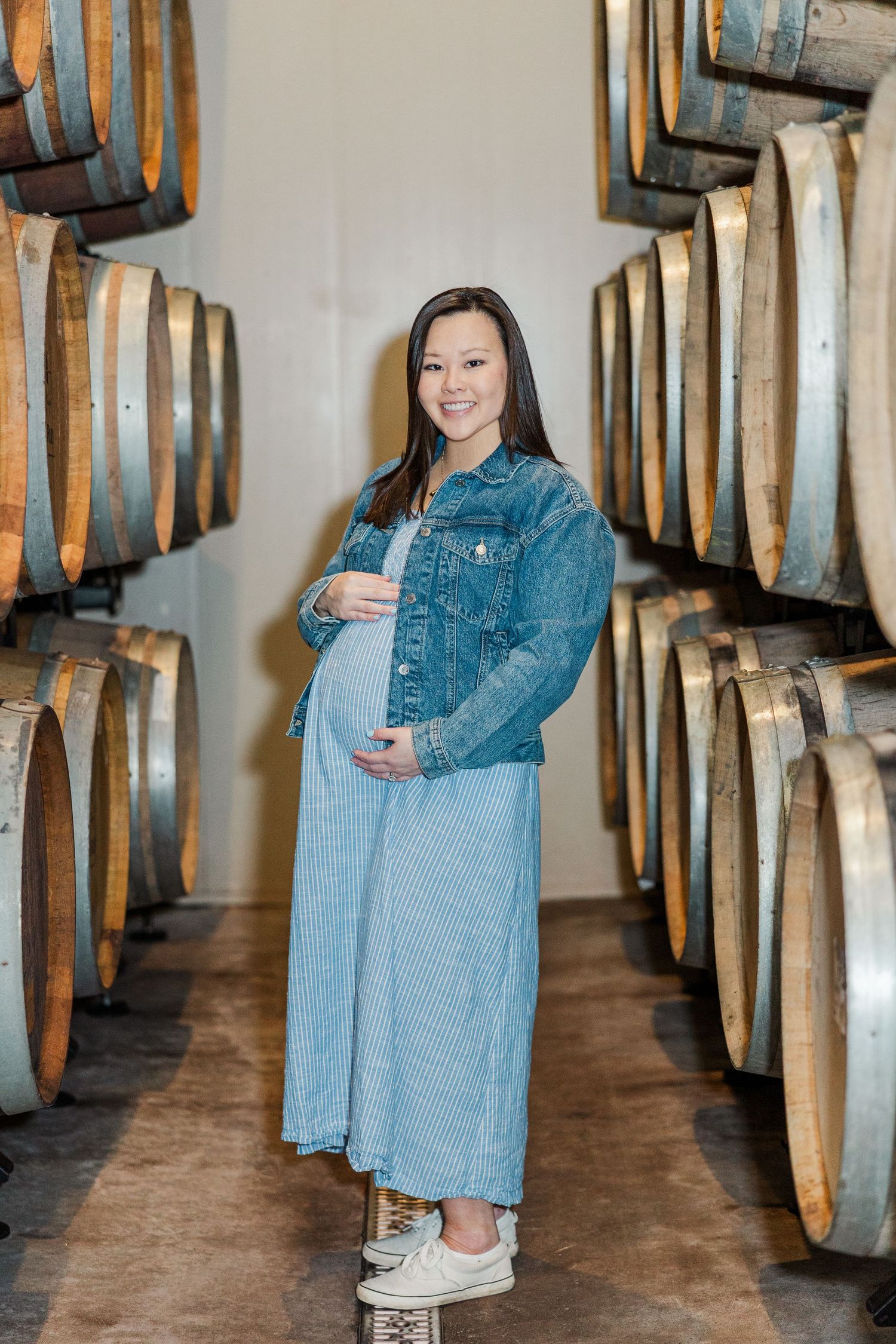 Pregnant woman in denim jacket and blue dress poses between oak wine barrels in a winery cellar.