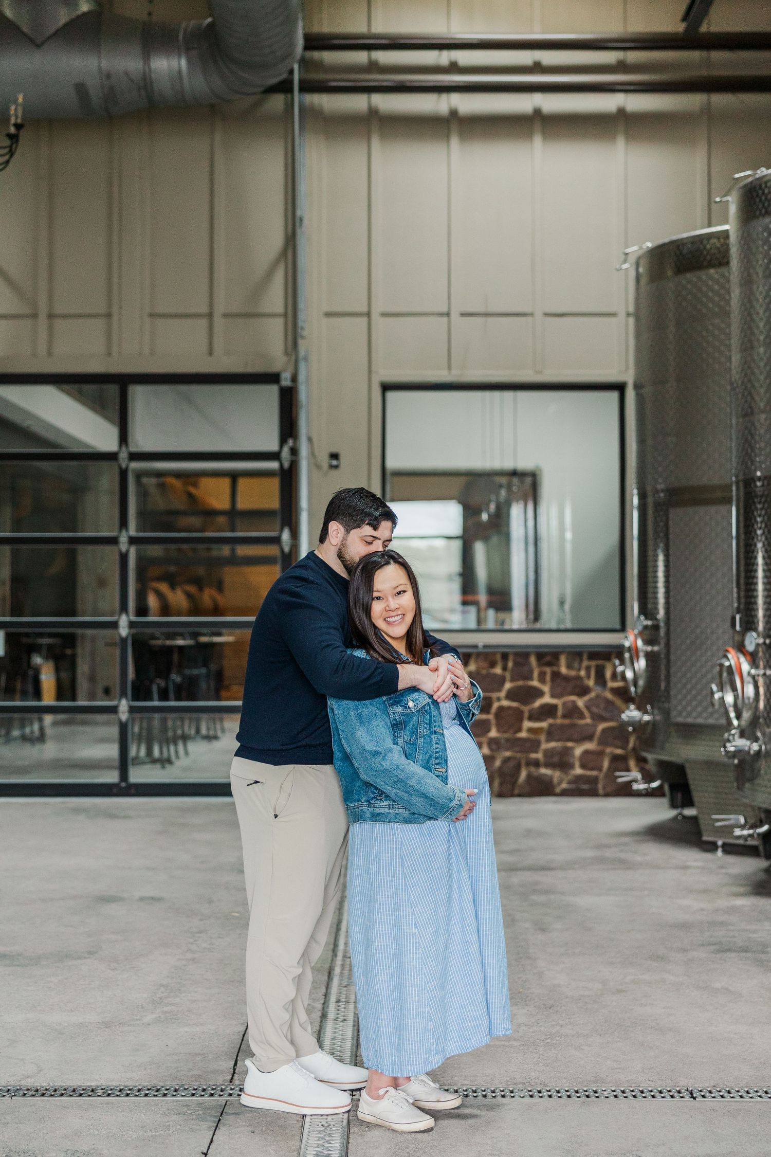 A couple embraces in a winery, the woman in a blue dress cradling her baby bump, surrounded by steel fermentation tanks.
