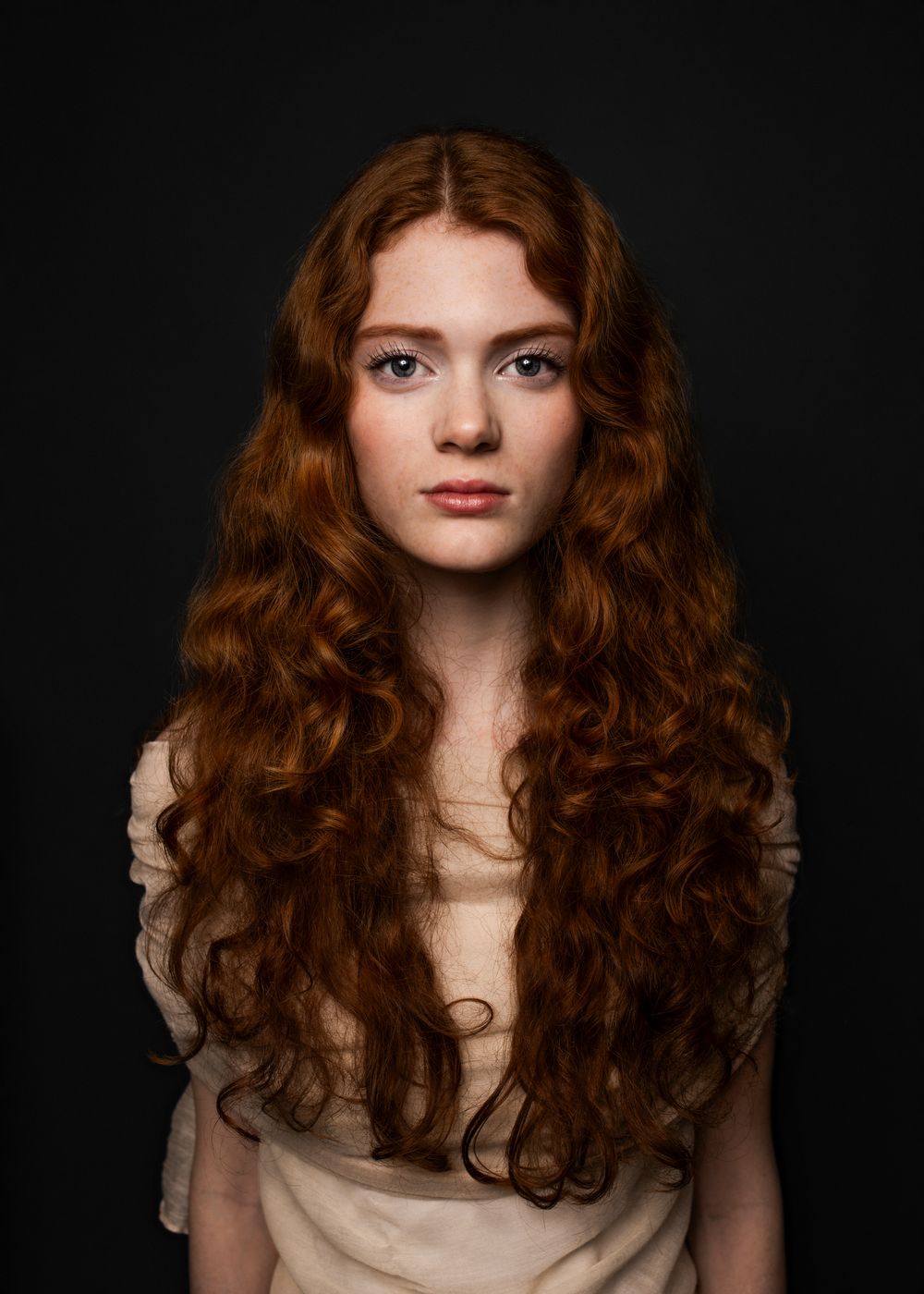 Young woman with long curly red hair and freckles posing against a dark background in a soft beige top.