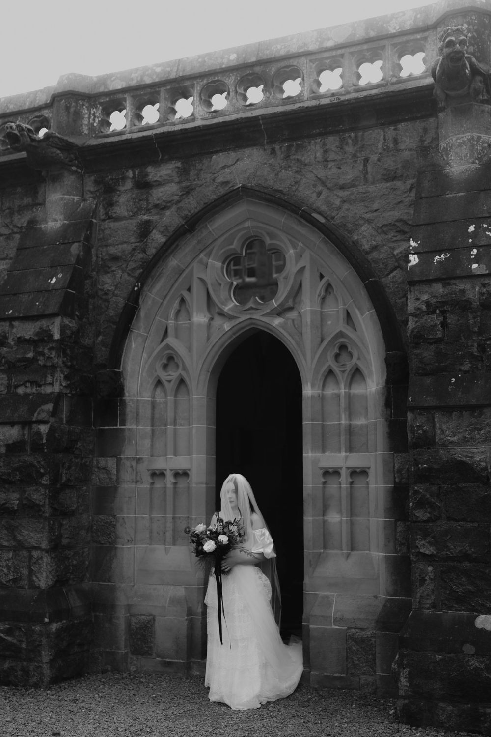Black and white photo of a bride in veil holding bouquet standing in Gothic church doorway.