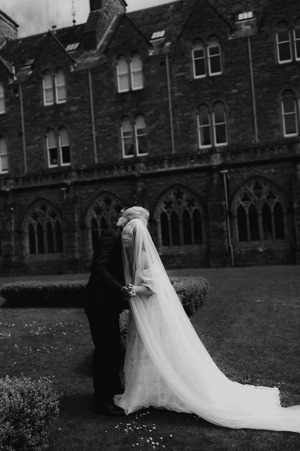 Black and white photo of a bride and groom kissing in front of a grand Gothic stone building on their wedding day.