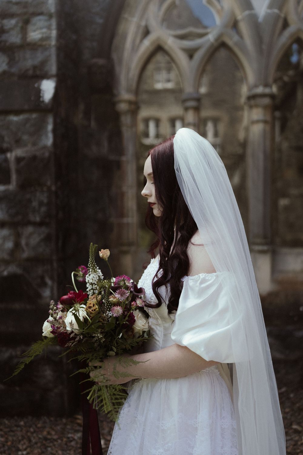 A bride in a white gown and veil holds a lush floral bouquet before a gothic stone church backdrop.