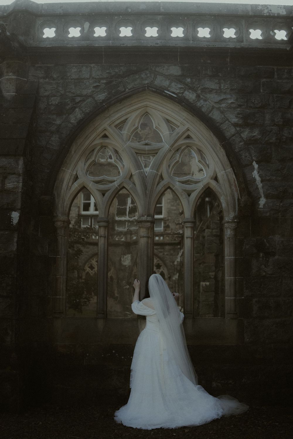 Bride in white gown and veil stands before ornate Gothic stone church doorway in moody, dramatic setting.