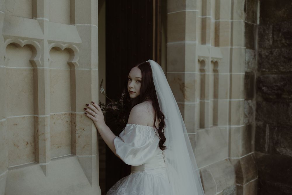A bride in an off-shoulder white gown and veil stands at a stone church doorway, her red hair flowing softly.