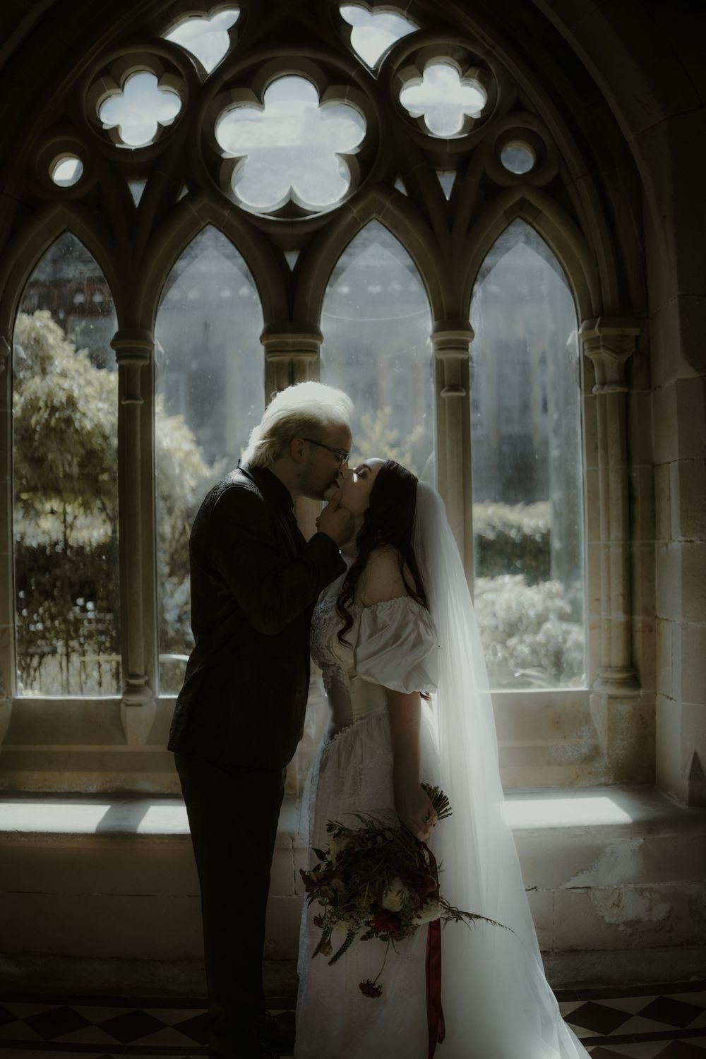 Bride and groom share a kiss in a Gothic cathedral archway, bride holding a bouquet in a white dress and veil.