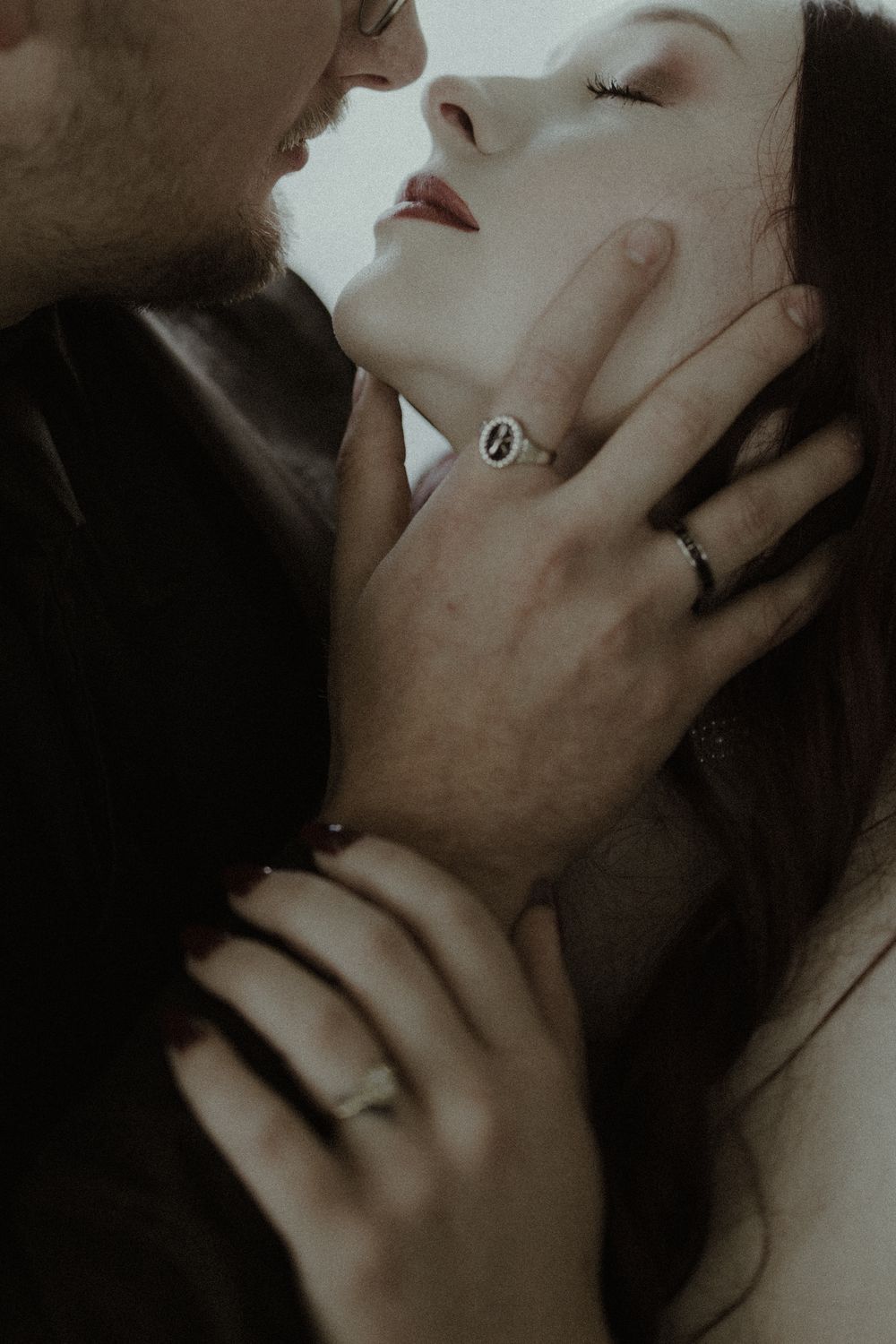 A romantic couple in an intimate embrace, man gently holding womans face, both wearing rings in moody dark tones.