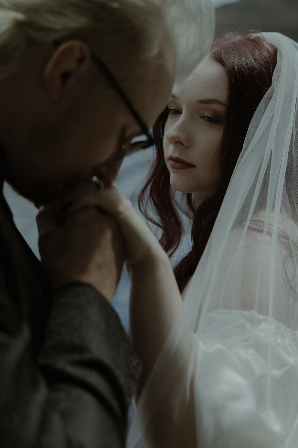 Groom in glasses kisses bride's hand as she wears a white veil and wedding dress in a romantic moody portrait.
