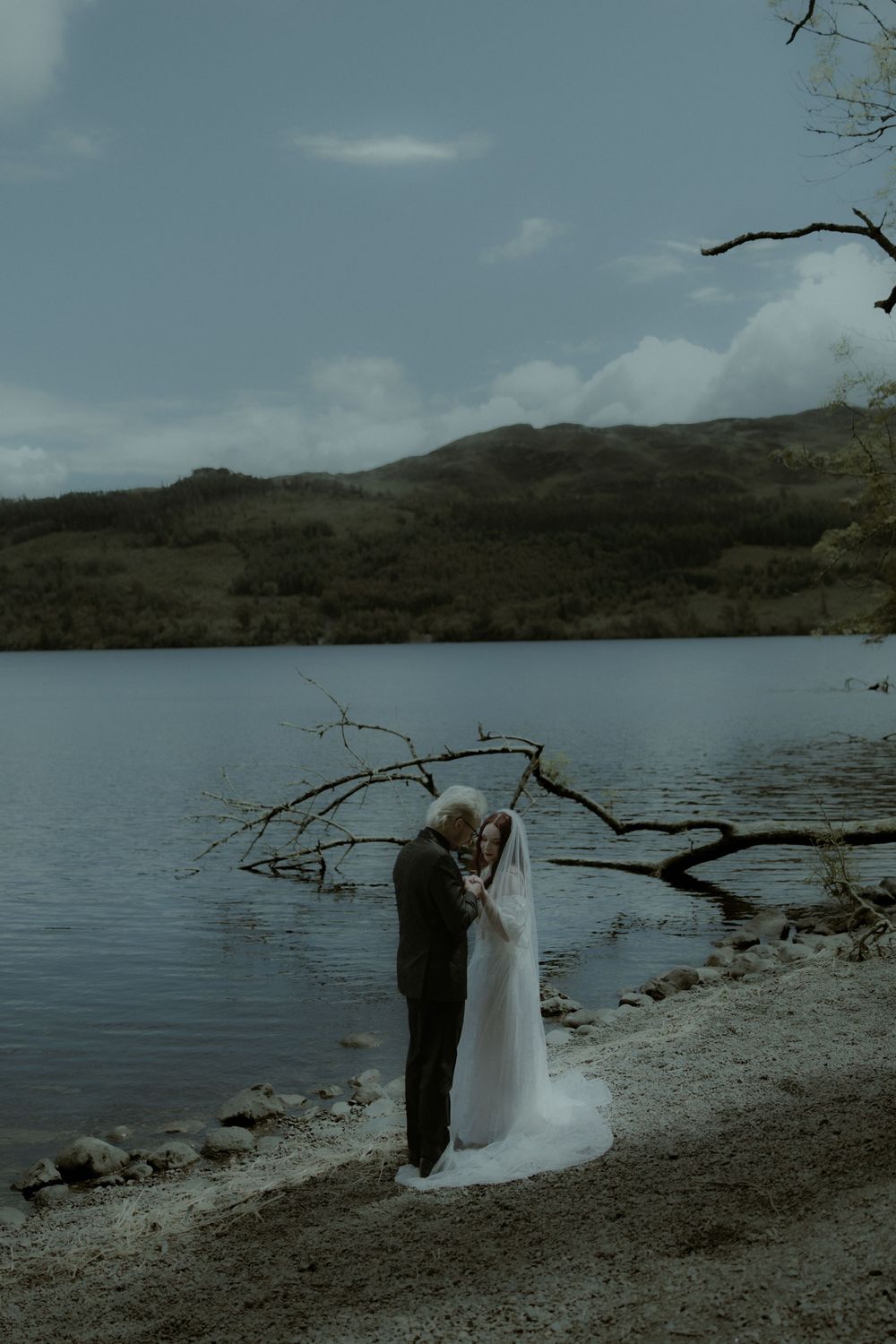 Bride and groom embrace by a serene lakeshore with mountains in the background on a moody, overcast wedding day.
