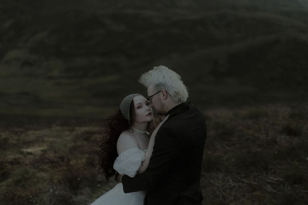 A couple shares an intimate moment in a moody highland landscape, the bride in white and groom in black embracing tenderly.