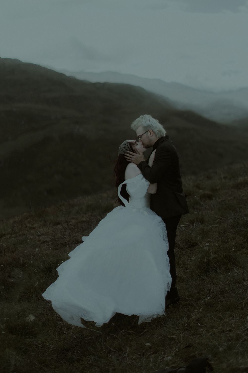 A couple shares a romantic kiss in wedding attire on a misty mountain hillside, bride in white gown, groom in dark suit.