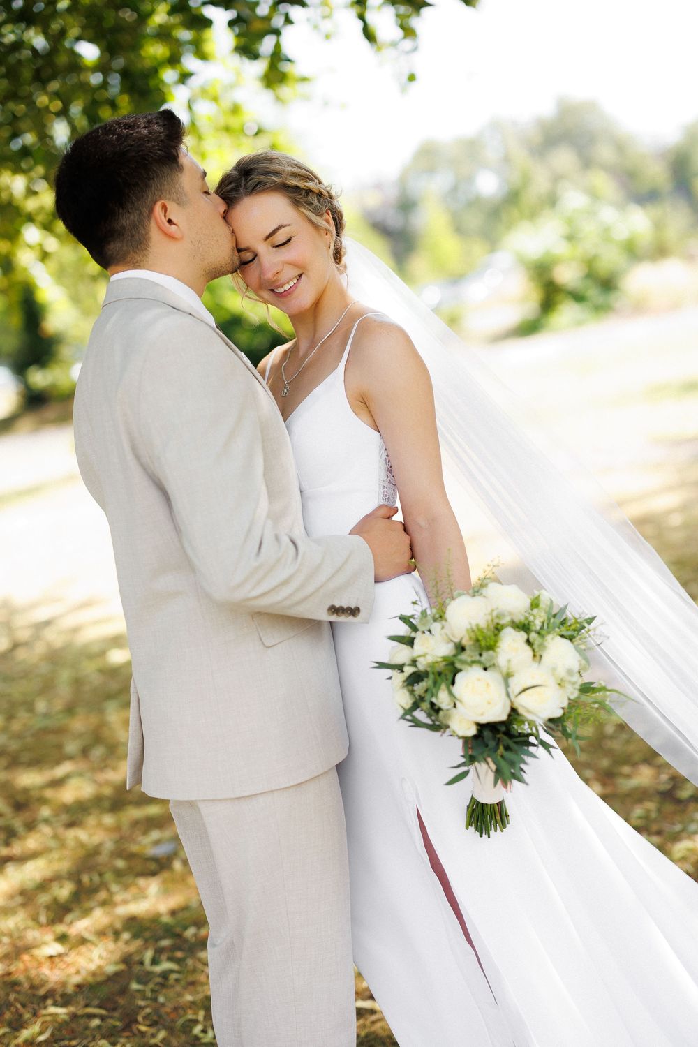 Groom kissing bride on cheek outdoors, bride holding white floral bouquet.