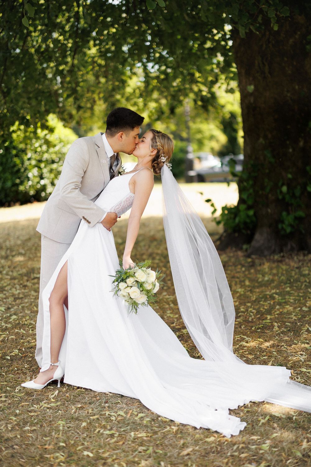 Bride and groom sharing a romantic kiss outdoors under a tree, bride holding white bouquet in flowing wedding dress.