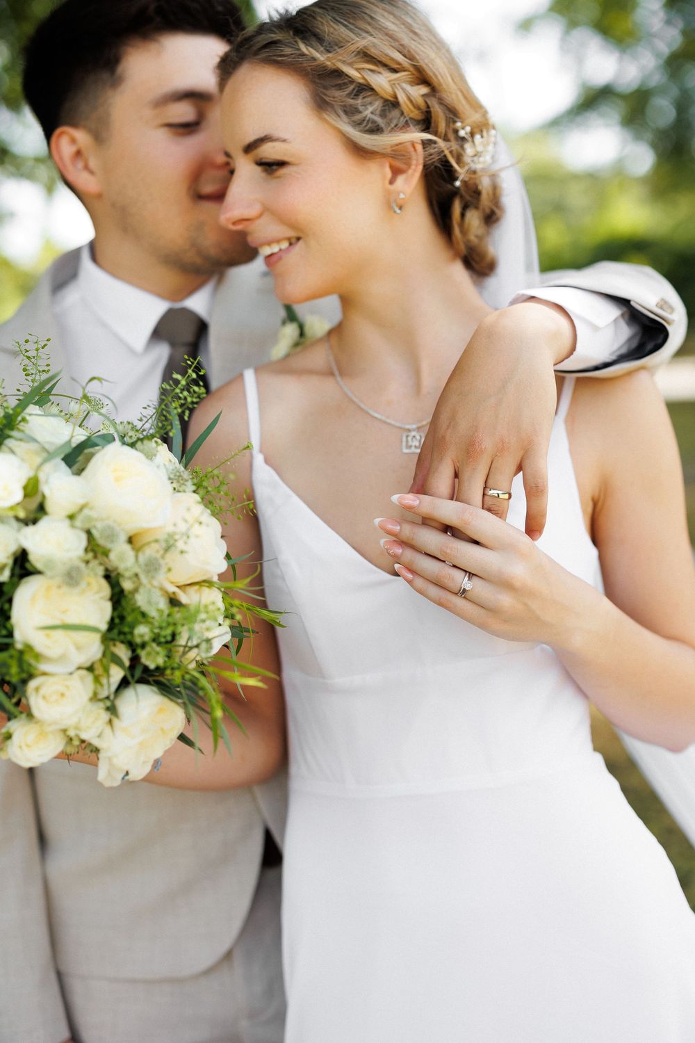 Happy bride in white dress holding bouquet while groom kisses her cheek outdoors.