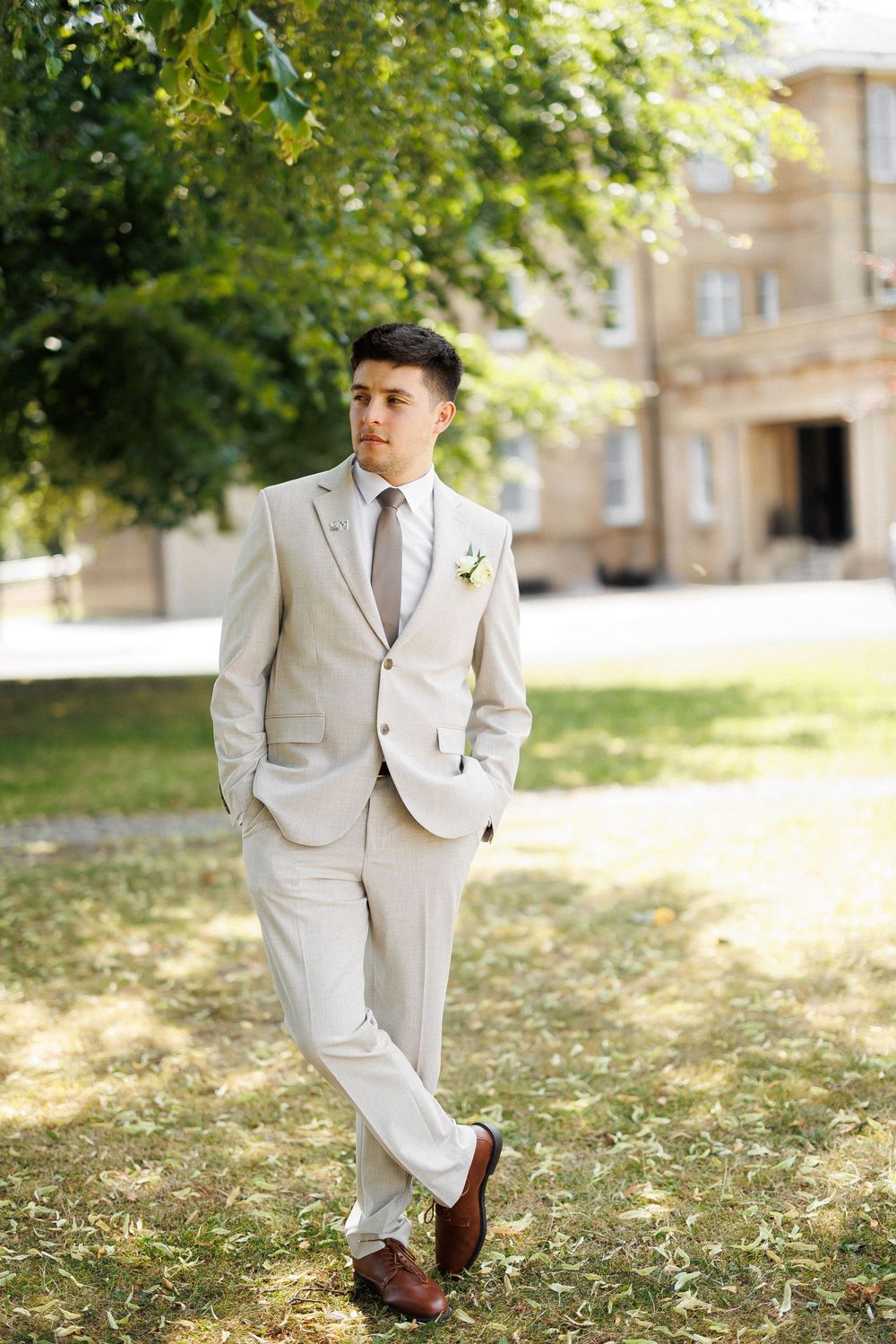 Stylish groom in beige suit with boutonniere standing outdoors in sunny garden near historic building.