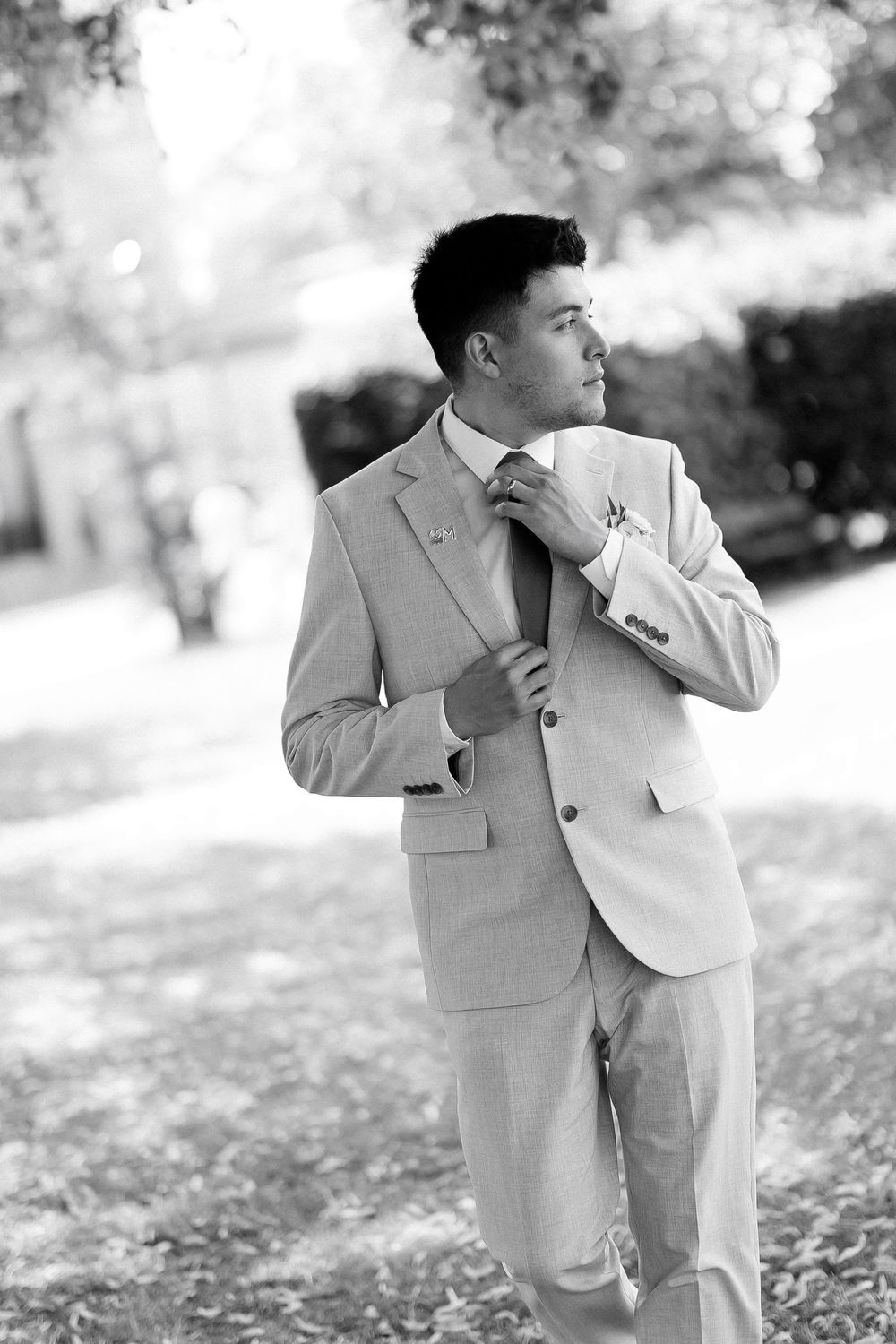 Elegant groom in light suit adjusting tie outdoors in black and white portrait photo.