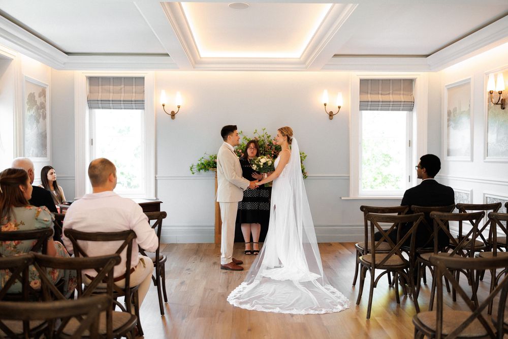 Bride and groom exchange vows at elegant indoor wedding ceremony with guests seated in wooden chairs.